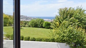 Une vue sur une ville côtière, prise depuis une fenêtre, qui inclut une zone herbeuse, des arbres et des bâtiments au loin.