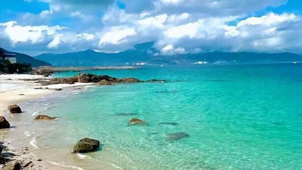 A beach with clear turquoise water, rocky shore, and distant mountains under a partly cloudy sky. - Zijijia Inn (Shenzhen)