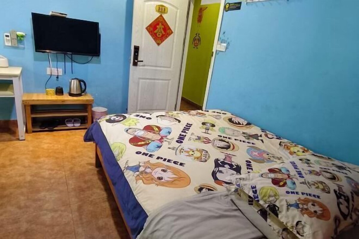 Un dormitorio con una cama con un edredón con temática de One Piece, un televisor montado en la pared, una mesita de noche de madera y una puerta decorada con un carácter chino rojo.