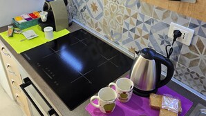 Keittiössä on musta induktioliesi, vedenkeitin ja kahvinkeitin. Purppuranvärisellä alustalla on kaksi mukea, viipale leipää ja lasi mehua.