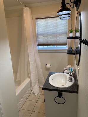 Ein Badezimmer mit einem weißen Waschbecken, einem Spiegel, einem Handtuchhalter, einem Fenster mit Jalousien und einer Duschvorhang.