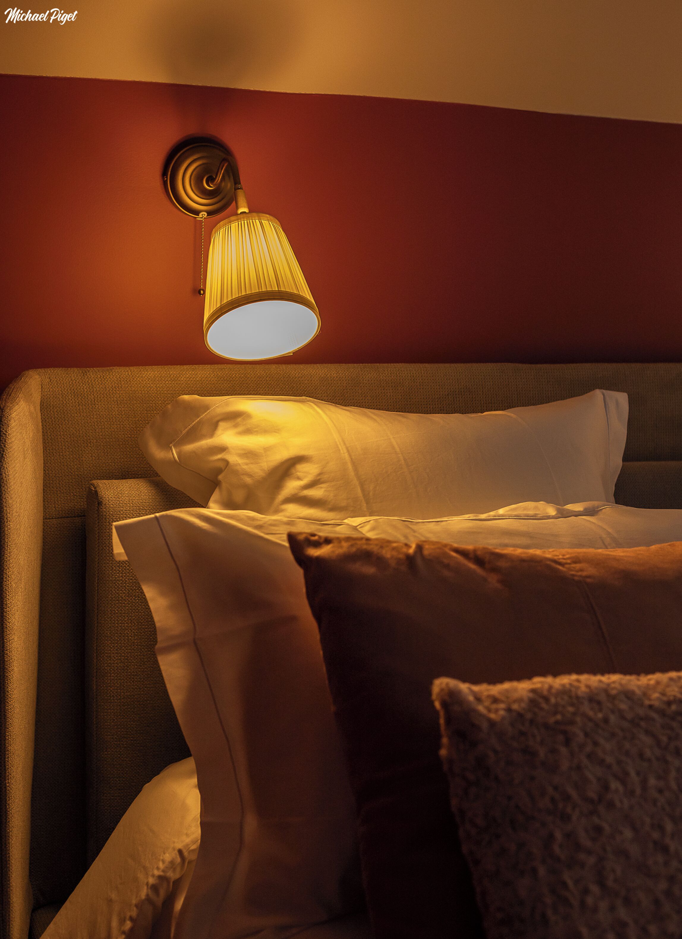 Quarto de hotel com cama, almofadas e uma luminária fixada na parede.