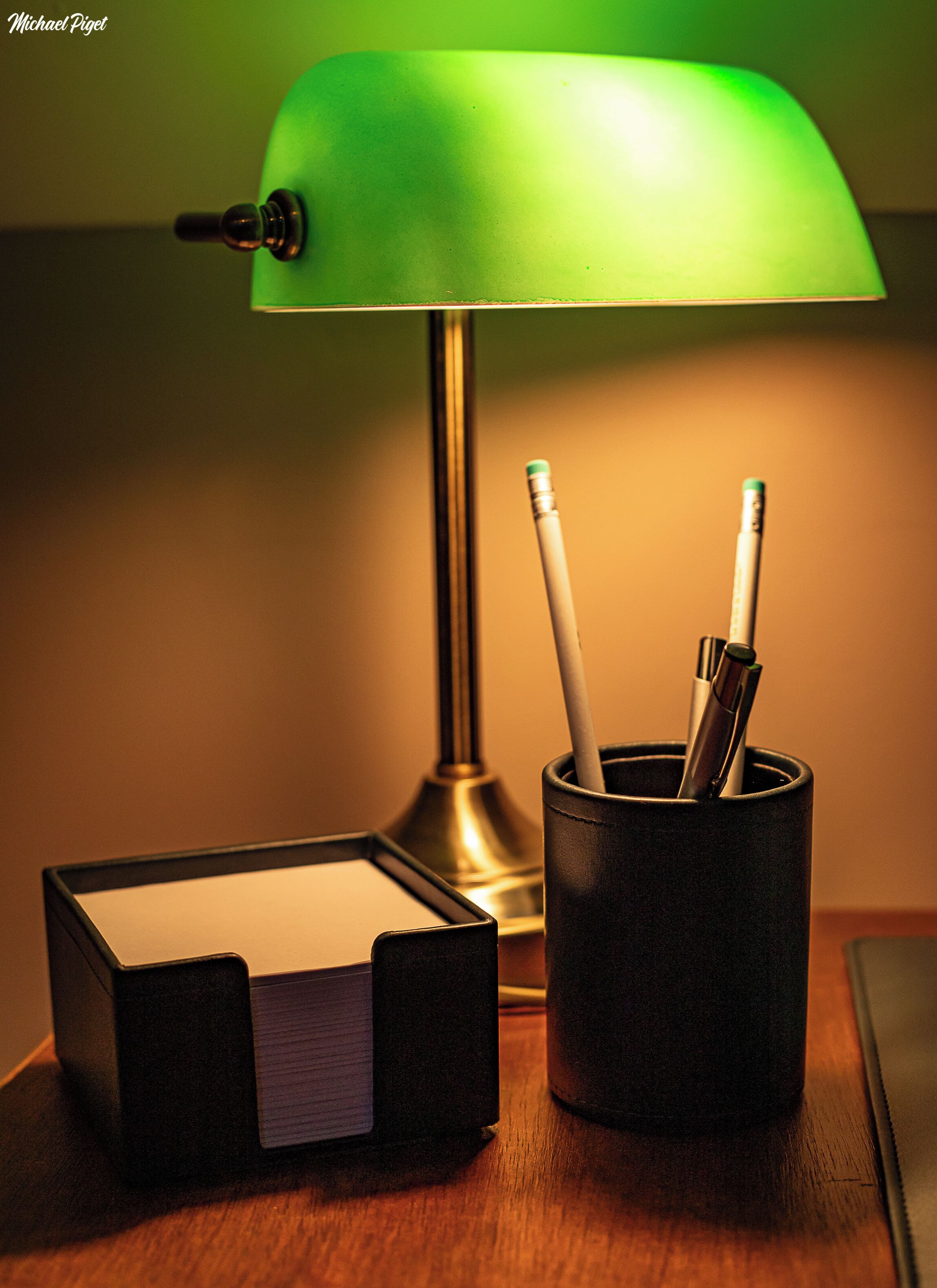 A desk with a green desk lamp, a black pen holder with pens, and a black notepad.