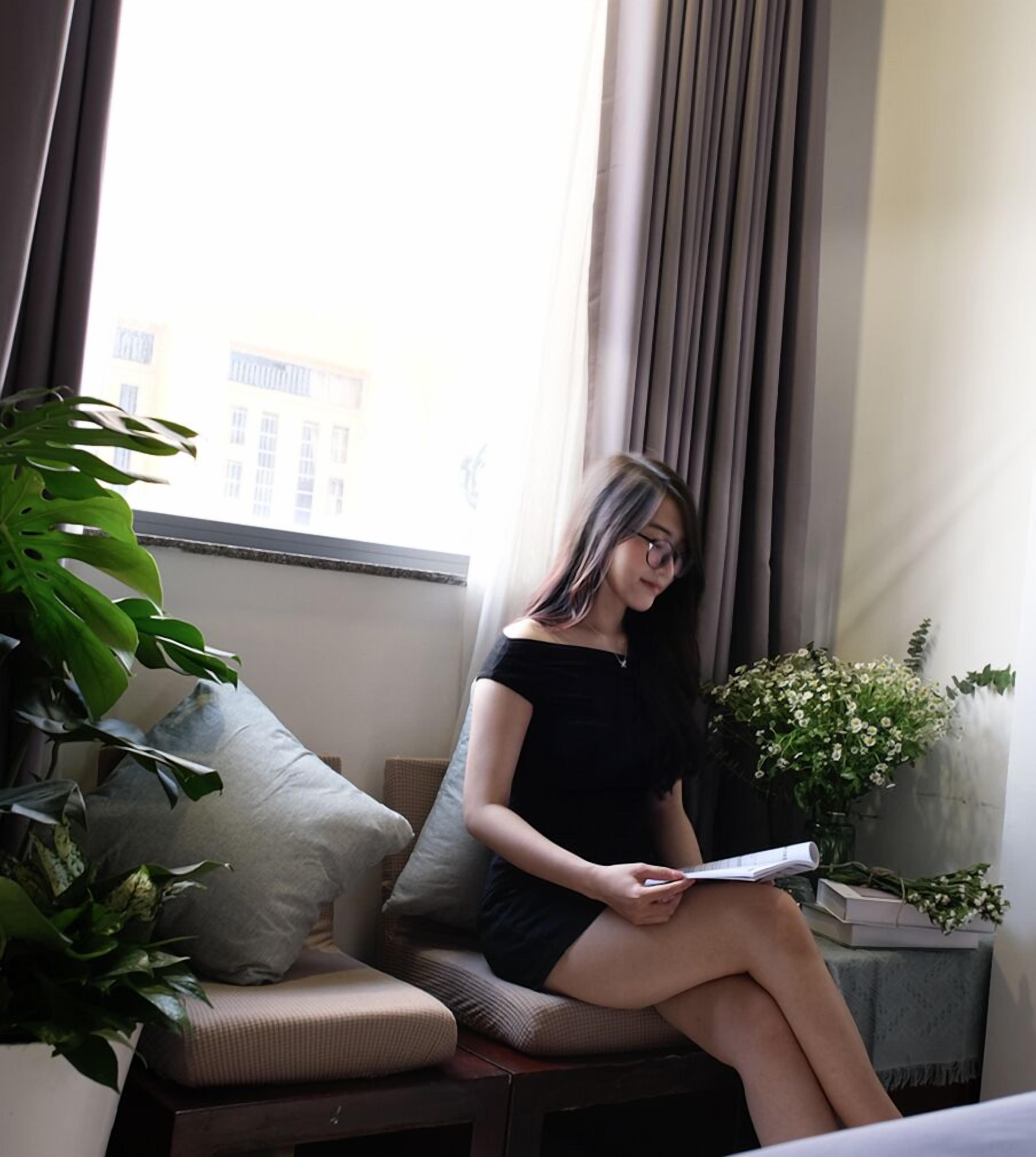 A person sitting on a sofa in a room with a window, a plant, and a vase with flowers.