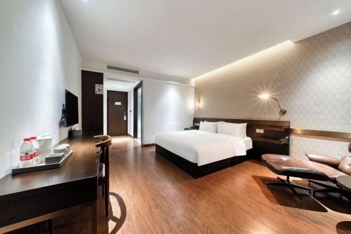 A modern hotel room with a large bed, a desk, a chair, and a desk lamp.