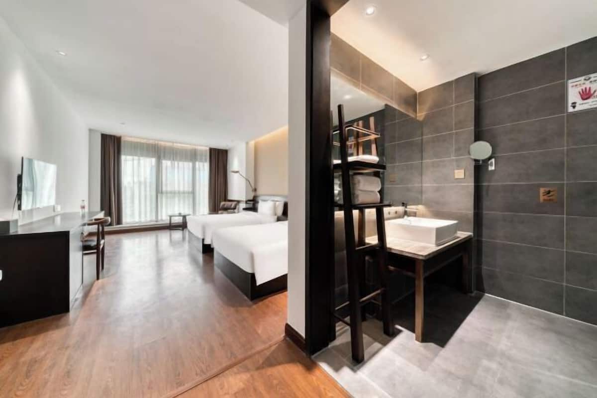 A hotel room with a wooden floor, a bed, a desk, a mirror, and a bathroom with a sink and towel rack.