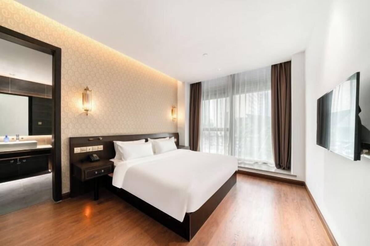 A modern hotel room with a large bed, a bedside table, a mirror, a TV, and a window with curtains.