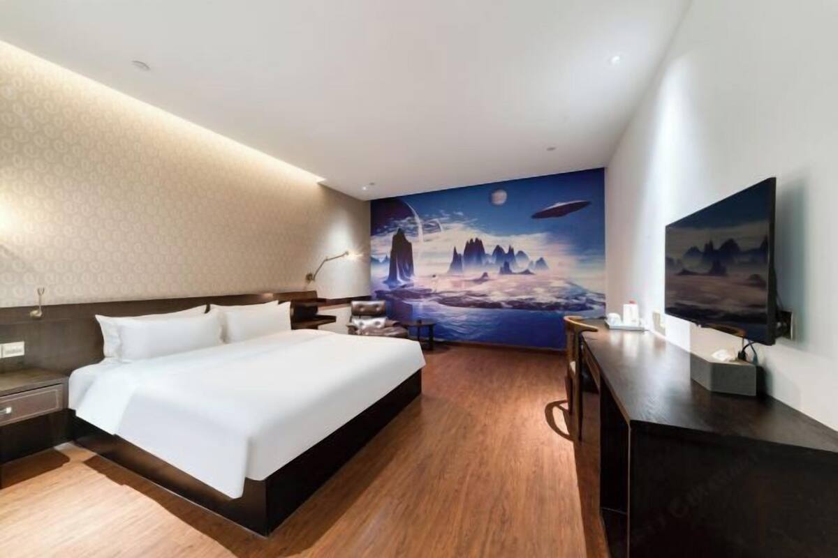 A hotel room with a large bed, a desk, a chair, a TV, and a mural on the wall.