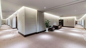 Un vaste couloir d’hôtel, doté d’un sol recouvert de moquette, d’un coin salon avec un canapé et des fauteuils, et d’une plante en pot.