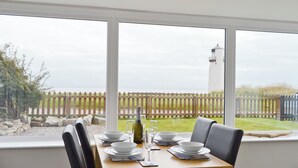 Ein Esstisch, gedeckt mit Tellern, Gläsern und einer Flasche, mit Blick auf einen Leuchtturm und einen Holzzaun.