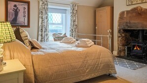 Ein Schlafzimmer mit Bett, Kamin, einem fensterbepflanzten Fenster, einem Sessel und einem Bild an der Wand.