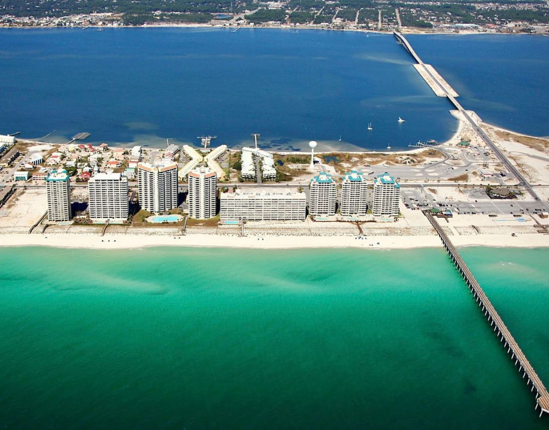 An aerial view of a coastal area with a long pier extending into the sea, surrounded by high-rise buildings and a sandy beach.