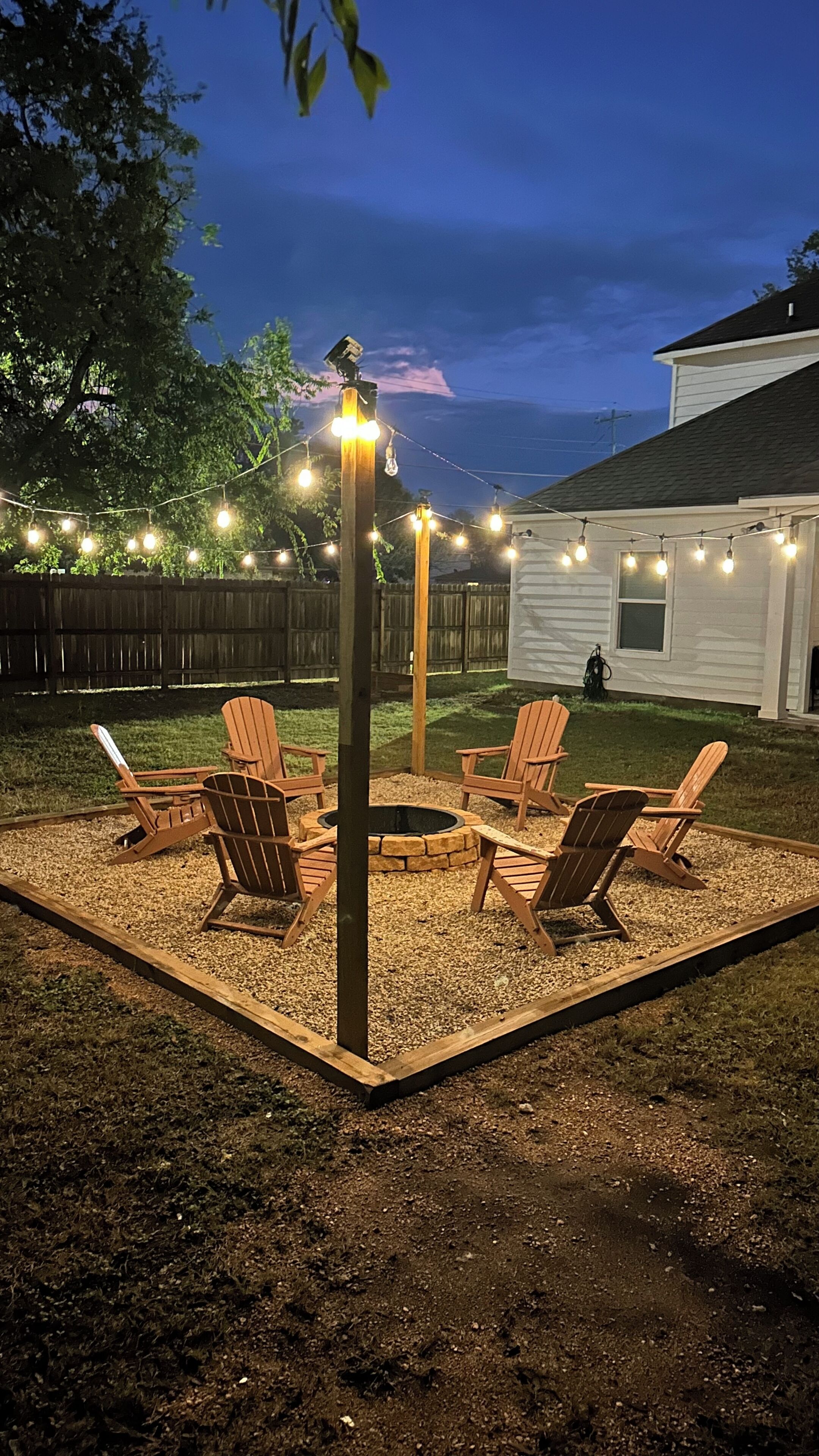 Un jardin avec un foyer entouré de fauteuils Adirondack, de guirlandes lumineuses et d’une terrasse en bois.