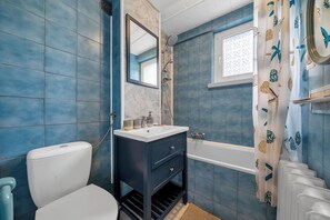 Bathroom features a white toilet, dark vanity, and bathtub with showerhead, set against blue-tiled walls and a decorative window.