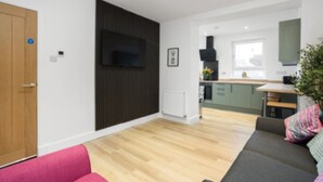 Ein modernes Wohnzimmer mit Holzboden, einem flachbildfernseher, der an einer dunklen Wand angebracht ist, einem rosafarbenen Sofa und einem grauen Sofa mit farbenfrohem Kissen. Im Hintergrund ist eine Küche mit grünen Schränken und einem Fenster erkennbar.