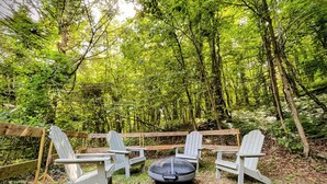 Eine Holzterrasse mit Adirondack-Stühlen und einer Feuerstelle in einem Waldgebiet.