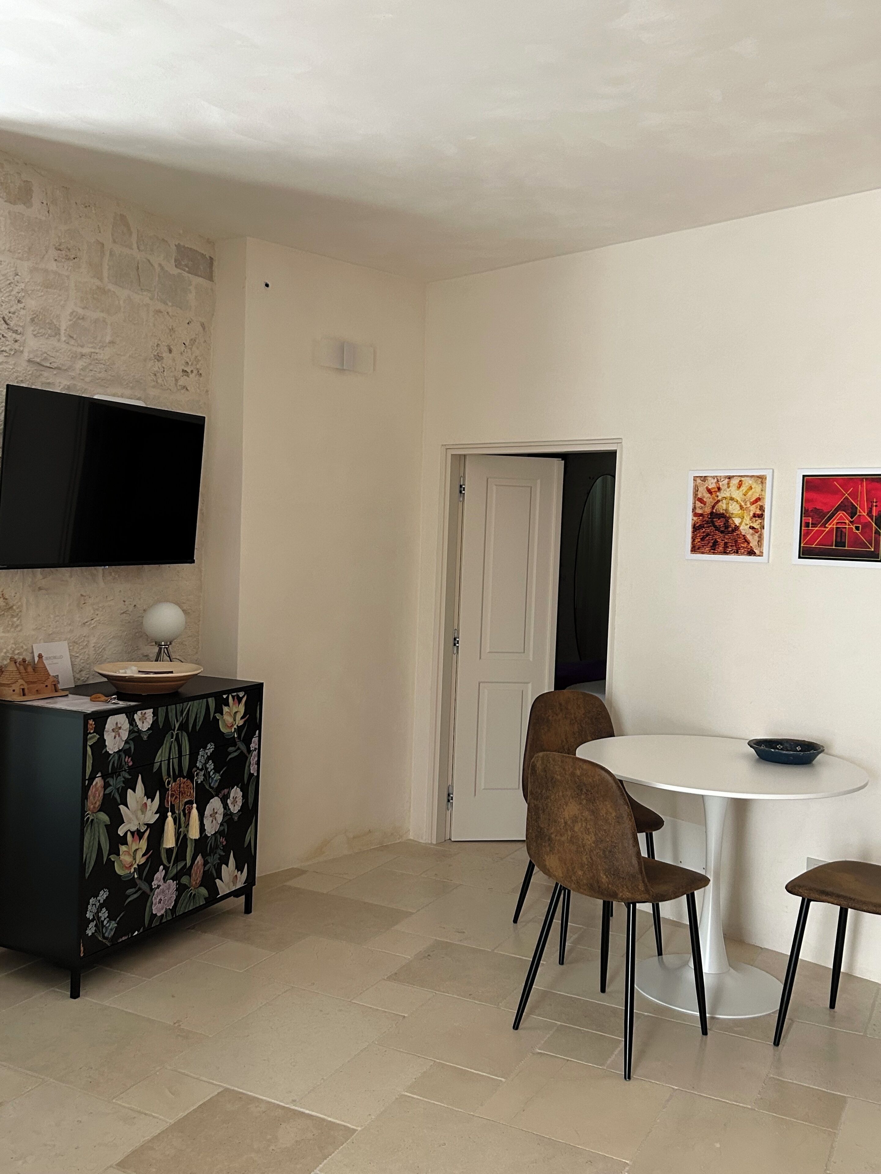 Un comedor con una mesa redonda, dos sillas, un mueble negro con motivos florales, un televisor de pantalla plana montado en la pared y dos obras de arte enmarcadas.