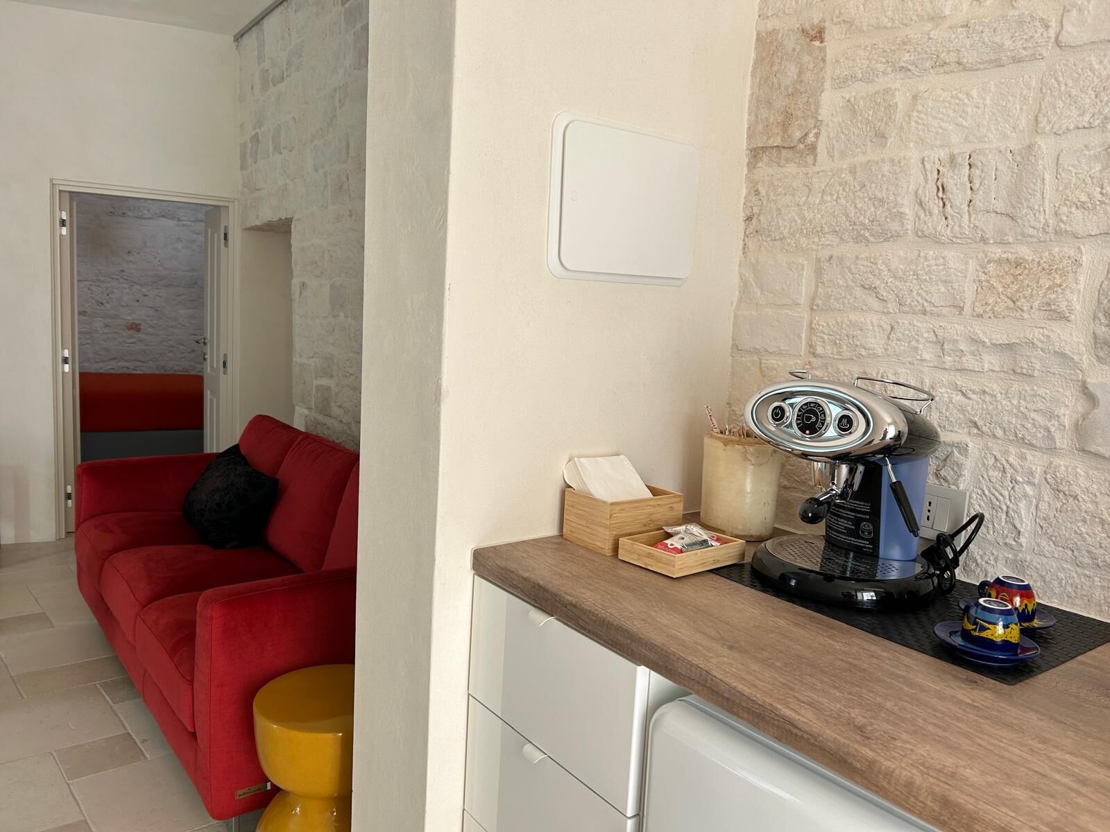 A compact kitchen with a coffee machine, a wooden side table, and a red sofa in the adjacent living area.