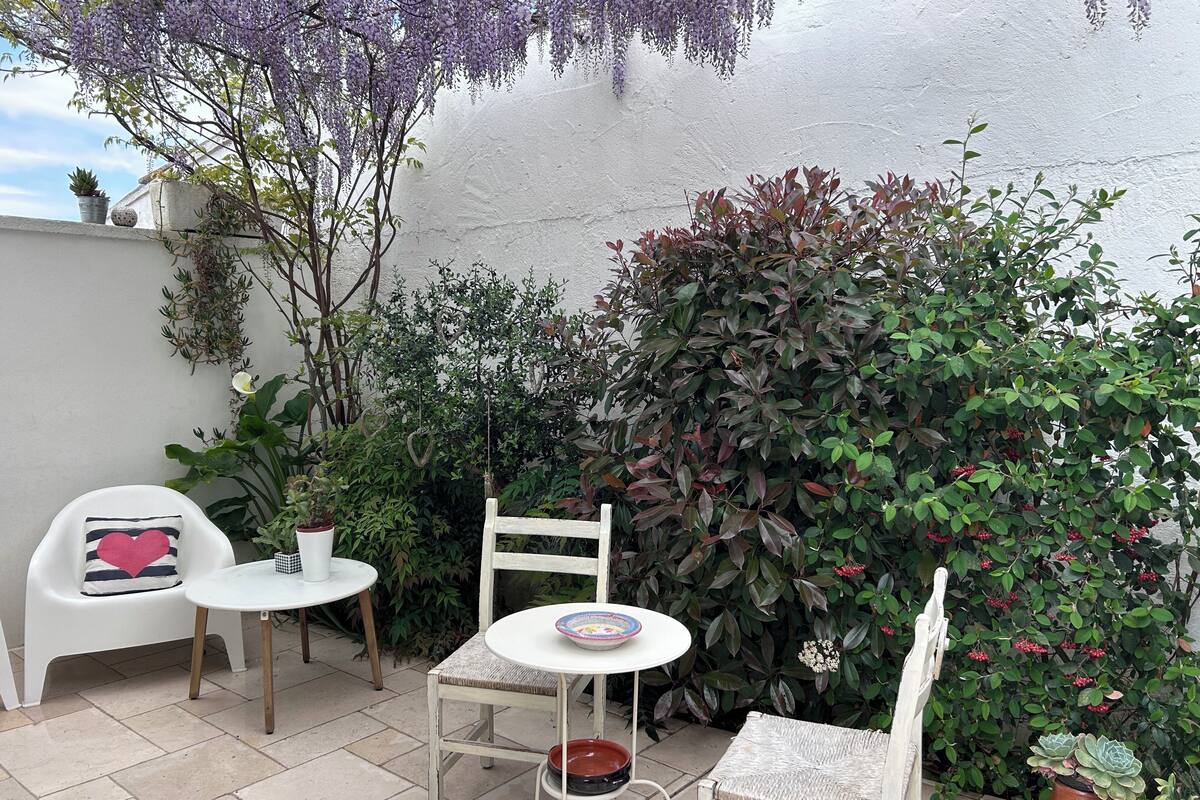 A small outdoor seating area with a white table and chairs, surrounded by various plants and a white wall.