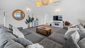 En moderne stue med grå sofaer, et træbord og et fjernsyn. Der er pendlerlamper i flet, et rundt spejl og en potteplante.