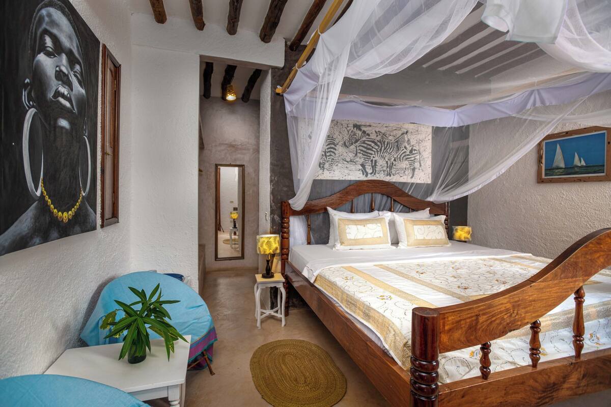 A bedroom with a wooden bed, a small bedside table, a nightstand, a potted plant, and a wall with a large painting.