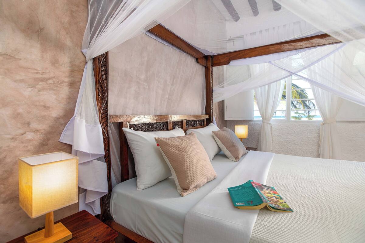 A bedroom with a canopy bed, white linens, and a bedside lamp.