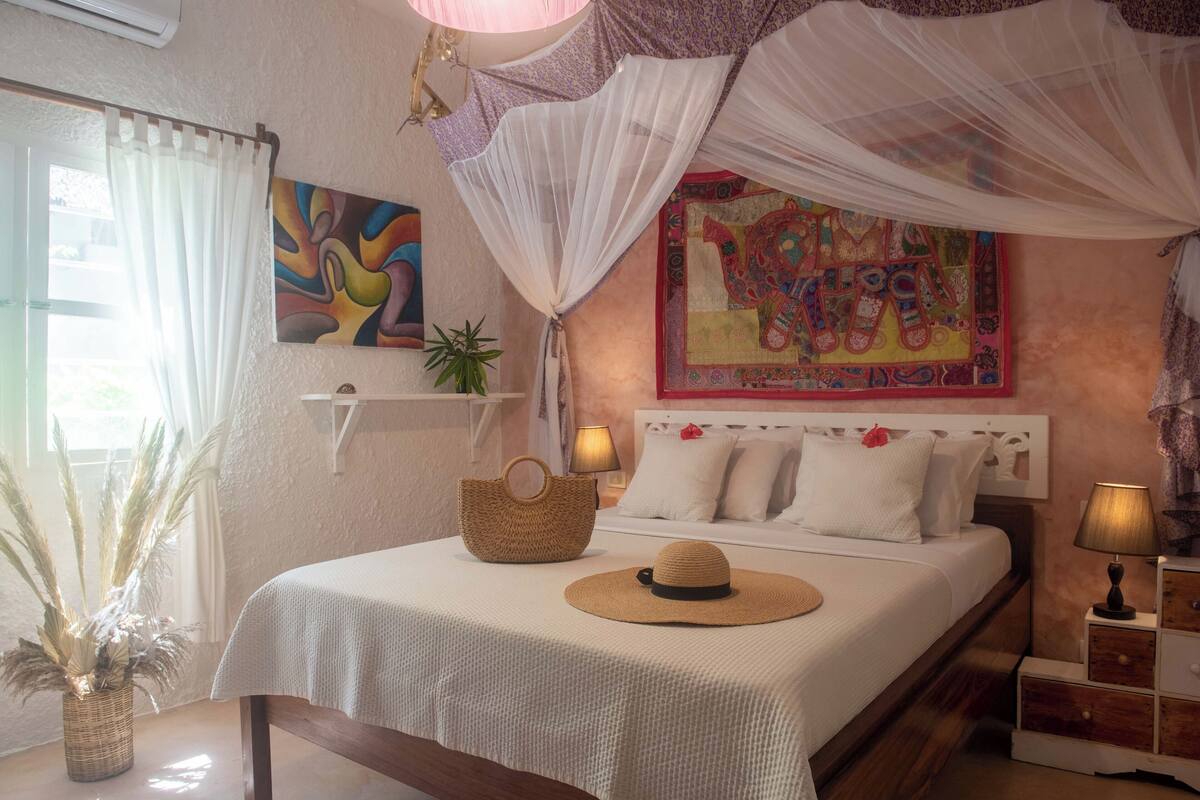 A neatly arranged bedroom with a bed, bedside table, lamp, decorative pillows, and a wall hanging.