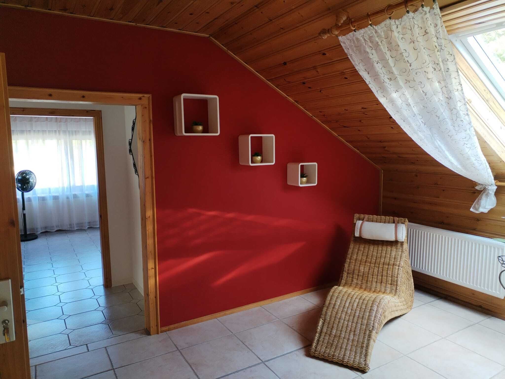Huoneessa on kalteva katto, punaiset seinät ja rottinkituoli. Seinällä on kolme pientä valkoista hyllyä, patteri ja ikkuna, jossa on valkoinen verho.
