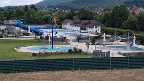 Ein Schwimmbeckenbereich mit Wasserrutsche, überdachten Sitzmöglichkeiten und Blick auf einen Berg im Hintergrund.