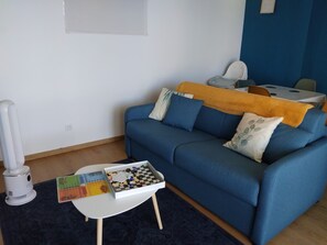 Ein Wohnzimmer mit einem blauen Sofa, einem kleinen weißen Tisch mit einem Brettspiel und einem weißen Gerät auf dem Boden.