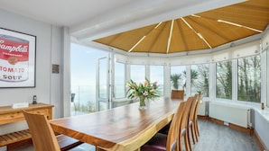 A spacious dining area with a large wooden table and chairs, a view of the sea through large windows, and a ceiling with a unique woven pattern.