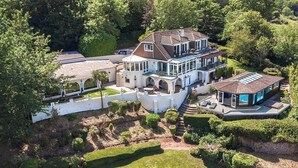 A large, two-story house with a spacious patio, surrounded by lush greenery and a white wall.