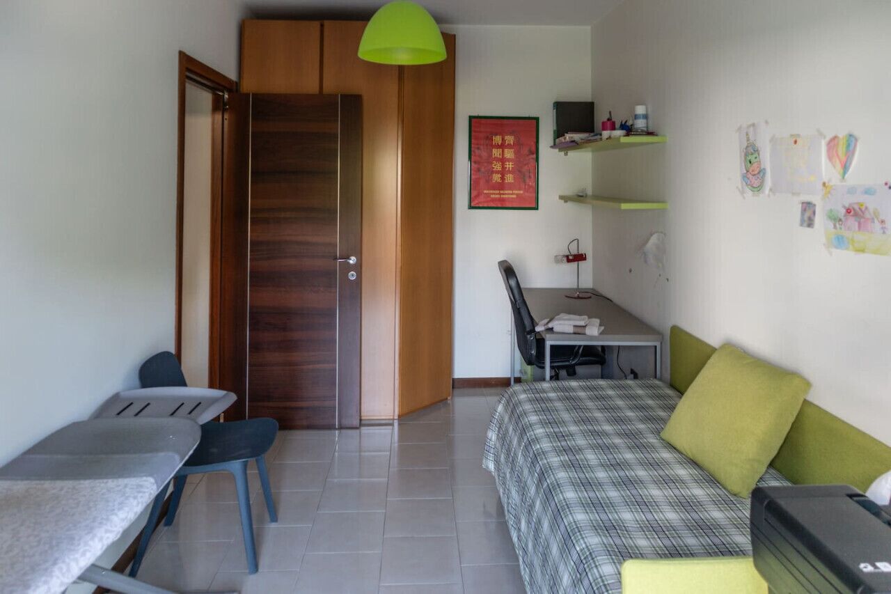 A compact room with a bed, a desk, and a chair. There's a wooden wardrobe, a green pendant light, and a red poster on the wall.