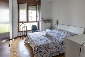 A bedroom with a wooden floor, a bed with a floral bedspread, a chair, a radiator, and a window with blinds. - Don Milani - Cernusco sul Naviglio (Cernusco sul Naviglio)