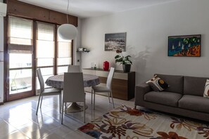 A modern living room with a sofa, a dining table with chairs, a coffee table, and a potted plant. There is a painting on the wall and a window with blinds. - Don Milani - Cernusco sul Naviglio (Cernusco sul Naviglio)