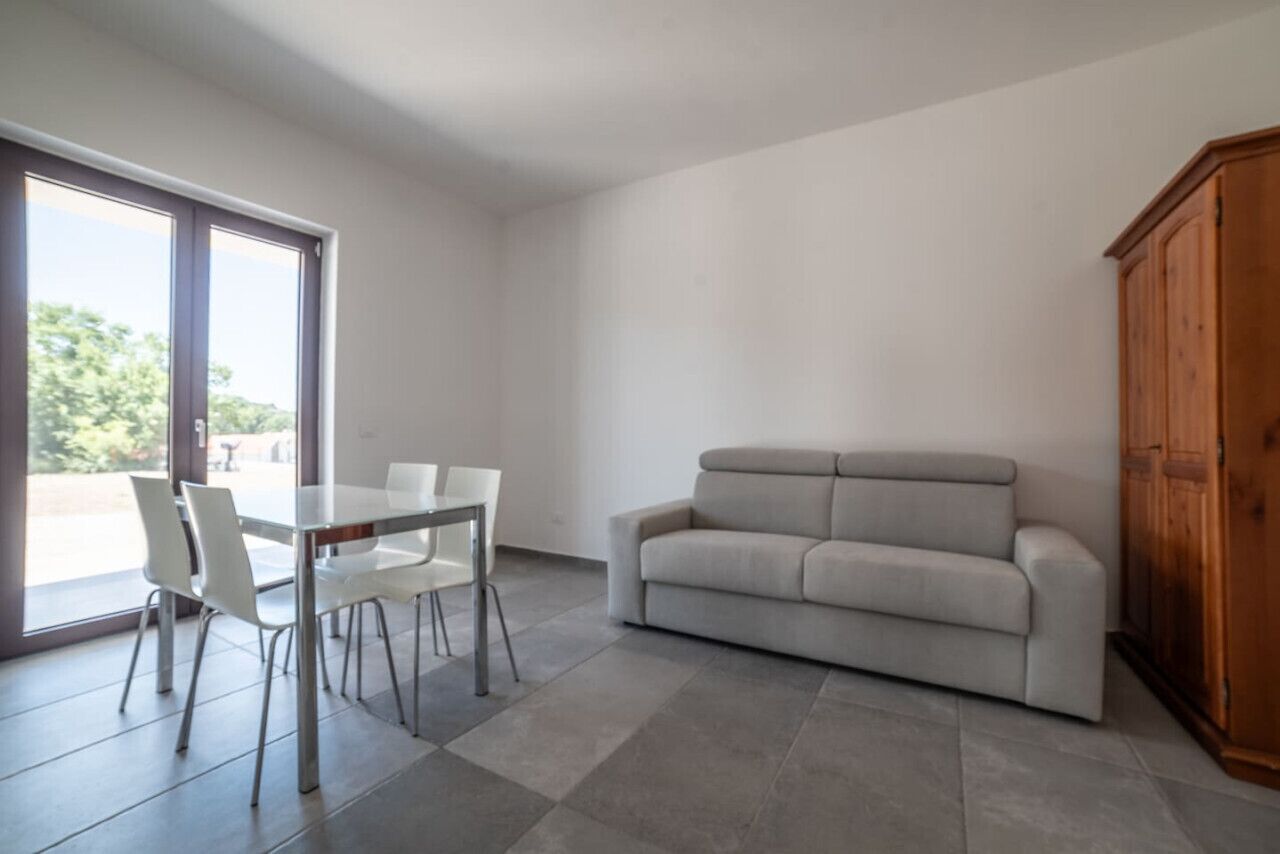 A modern living room with a glass dining table, white chairs, a grey sofa, and a wooden cabinet.