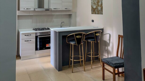 Une cuisine moderne avec des meubles blancs, un îlot central doté de tabourets de bar et un coin repas avec une chaise en bois.