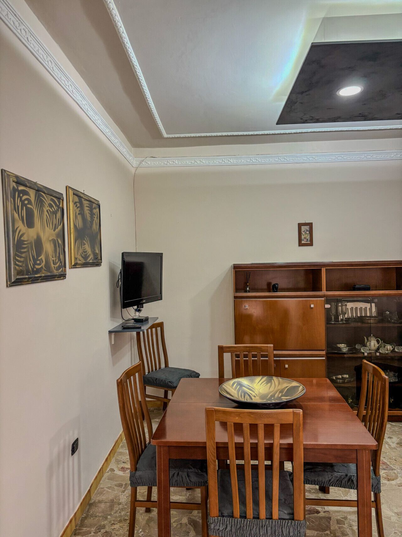 A dining area with a wooden table and chairs, a television on a stand, and a decorative cabinet with glass doors.