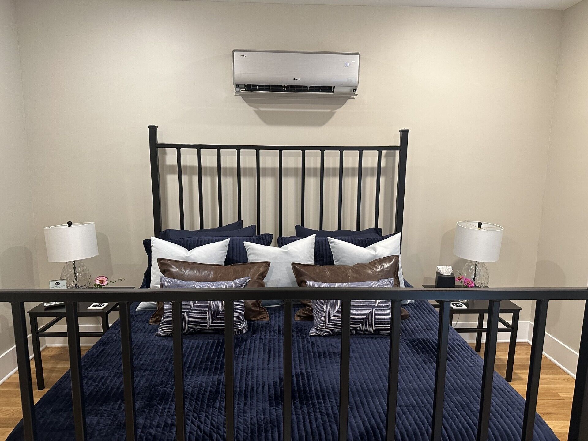 Une chambre avec un lit en métal noir, une couvre-lit matelassée bleu foncé et de nombreux coussins aux motifs et couleurs variés. Un appareil de climatisation est fixé au mur au-dessus du lit.