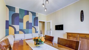 A dining area with a wooden table, chairs, a vase of flowers, a colorful abstract wall, a television, and a wooden cabinet. - The Popes' Oasis - 2 steps from Trastevere and Historical Center (Roma)