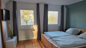 Une chambre avec un lit en bois, deux fenêtres avec des rideaux, un radiateur et un téléviseur fixé au mur.