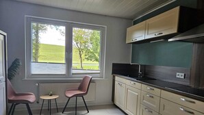 Une cuisine compacte donnant sur un champ verdoyant, avec deux chaises, une petite table et des armoires de couleur beige.
