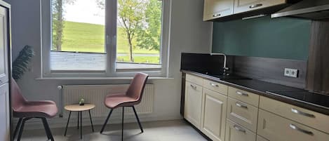 Uma cozinha compacta com vista para um campo verde, duas cadeiras, uma mesa pequena e armários bege.