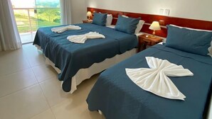 Een hotelkamer met twee bedden, elk voorzien van een blauwe sprei en witte handdoeken. Er is een plafondventilator, een nachtkastje met een lamp, en uitzicht op de omgeving door een groot raam.