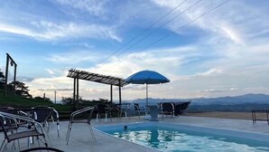 Een zwembadterras met ligbedden en parasols, een heldere lucht en bergen op de achtergrond.