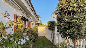 A narrow, paved pathway flanked by potted plants and a white fence, leading to a building with a tiled roof and a visible door.