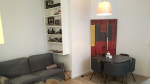 Una sala de estar con un sofá gris, una mesa de centro de vidrio, una mesa de comedor con cuatro sillas, y una estantería en la pared con libros y adornos.