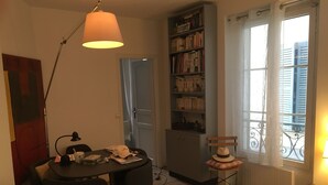 Una habitación pequeña y acogedora con una mesa de comedor, sillas, una estantería, un radiador y una ventana con vistas a edificios.