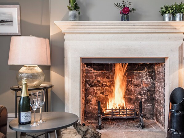 A fireplace with a log fire, a table with a bottle and glass, and a lamp.