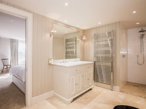 A modern bathroom with a large mirror, a double sink vanity, a walk-in shower, and a bed in the adjacent room.